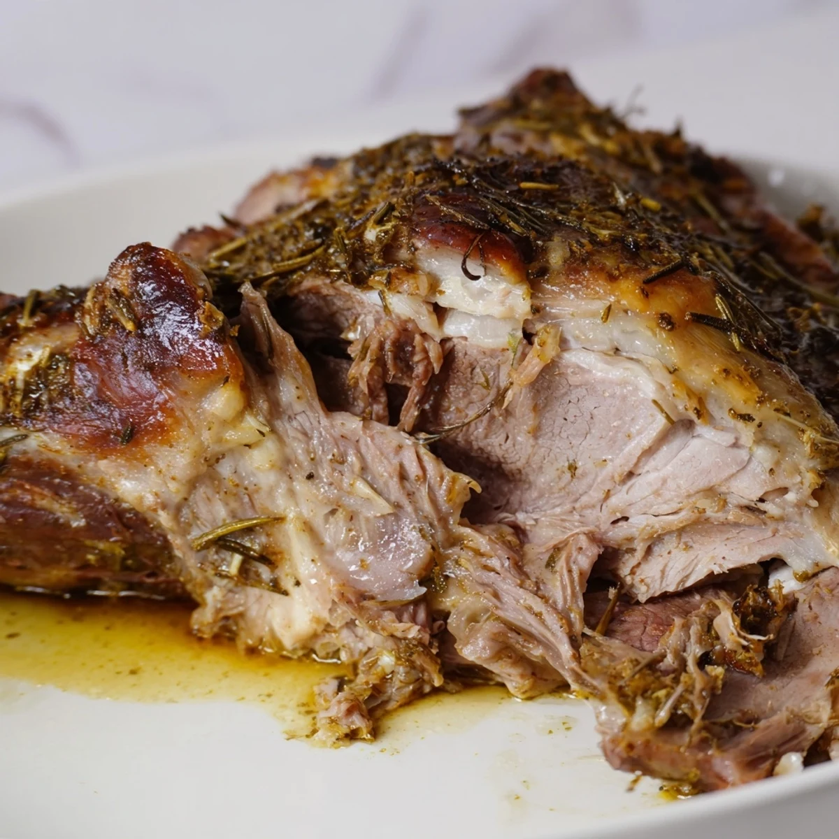 Fresh rosemary sprigs and garlic cloves rest beside a sliced, juicy portion of slow roasted lamb shoulder with herbs.