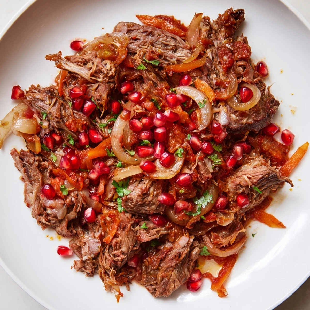 An overhead view of Braised Lamb Shoulder with Pomegranate shows the succulent meat, vibrant seeds, and a drizzle of rich, aromatic sauce.