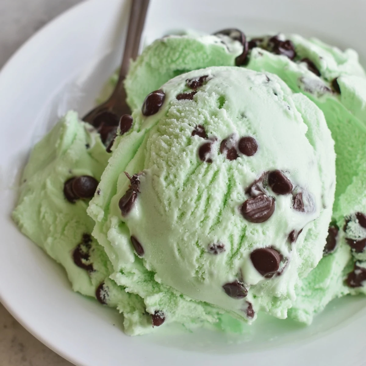 Green mint chocolate chip ice cream in a white bowl with fresh mint leaves on a marble counter.