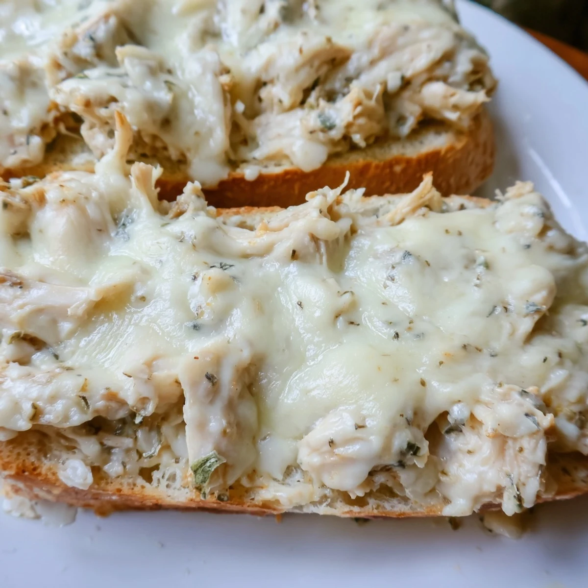 A close-up of viral chicken Alfredo garlic bread shows melted cheese over a hearty chicken Alfredo spread on toasted bread.