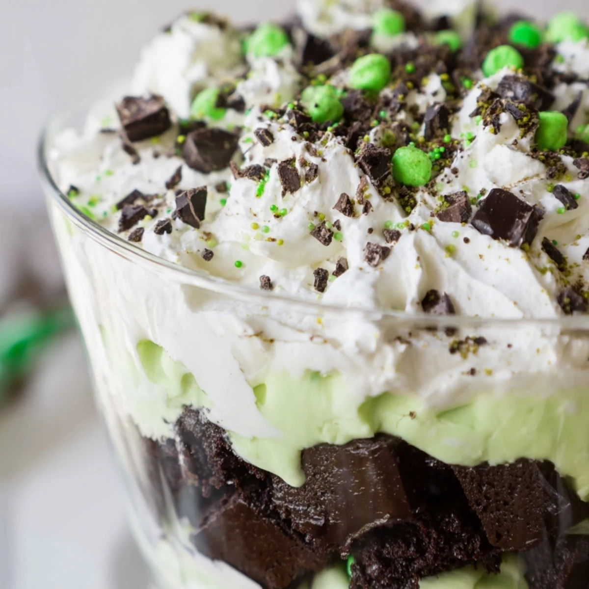 Glass trifle bowl showcasing layered St. Patricks Day brownie trifle with chocolate brownie cubes, green pistachio pudding, and fluffy whipped cream topped with green sprinkles