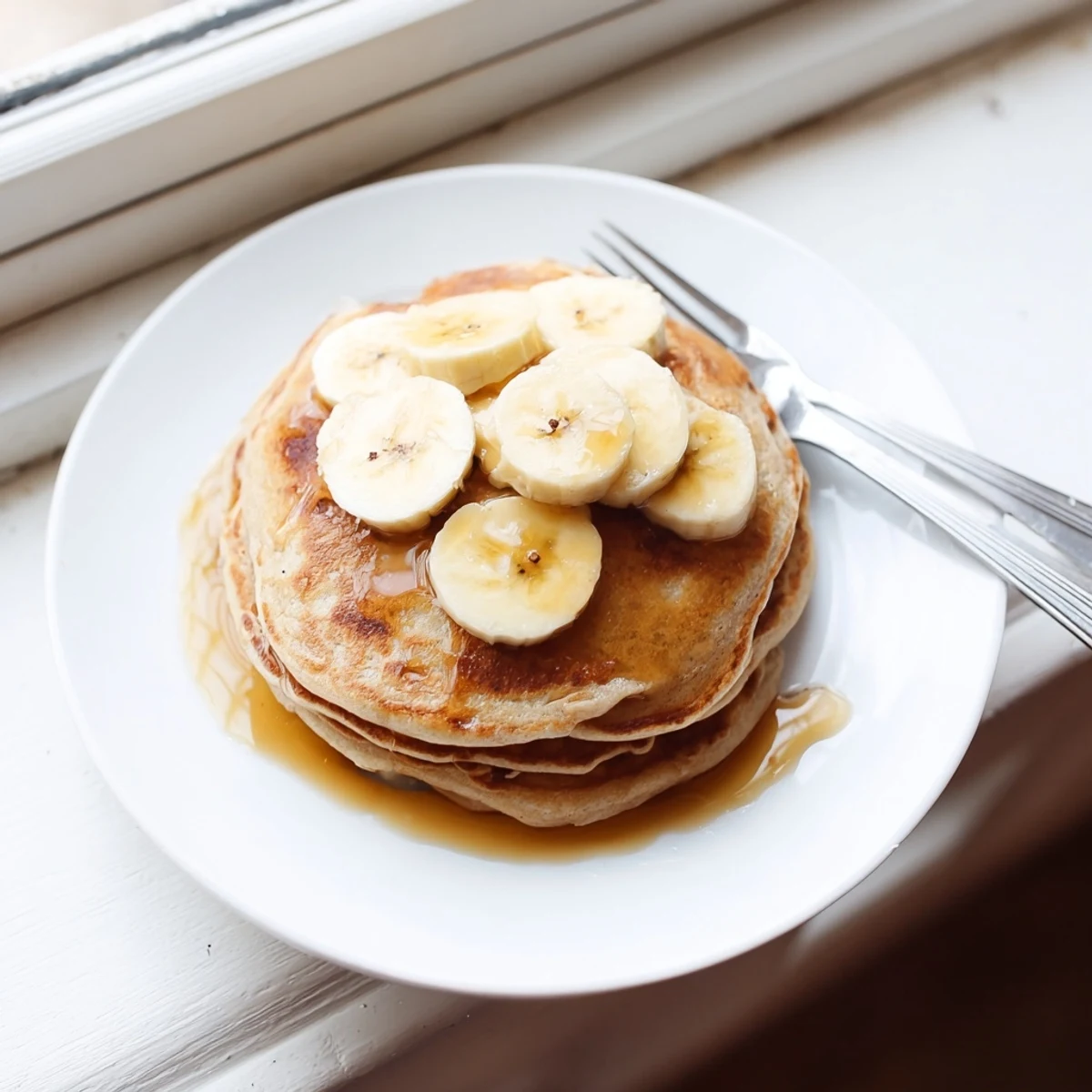 Golden stack of fluffy Greek yogurt banana pancakes drizzled with maple syrup and topped with fresh banana slices.